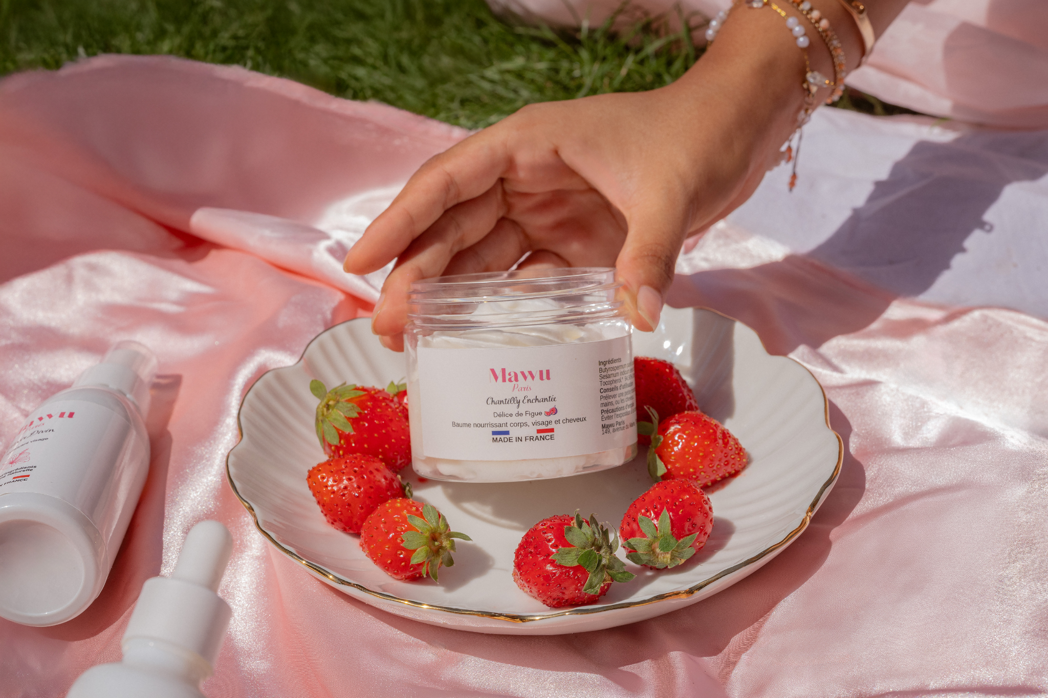 Un femme à la peau mate tient un peau de Chantilly Enchantée dans une assiette de porcelaine entourée de fraises. L'assiette est posé sur une nappe en satin rose sur l'herbe.
