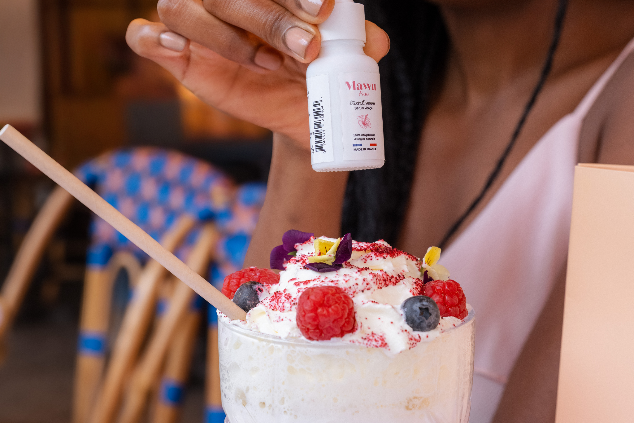 Une jolie femme noire tient de sa main droite un flacon de sérum Élixir D'amour au dessus d'un milkshake  rose, avec de la chantilly au -dessus des framboises et des myrtilles.
