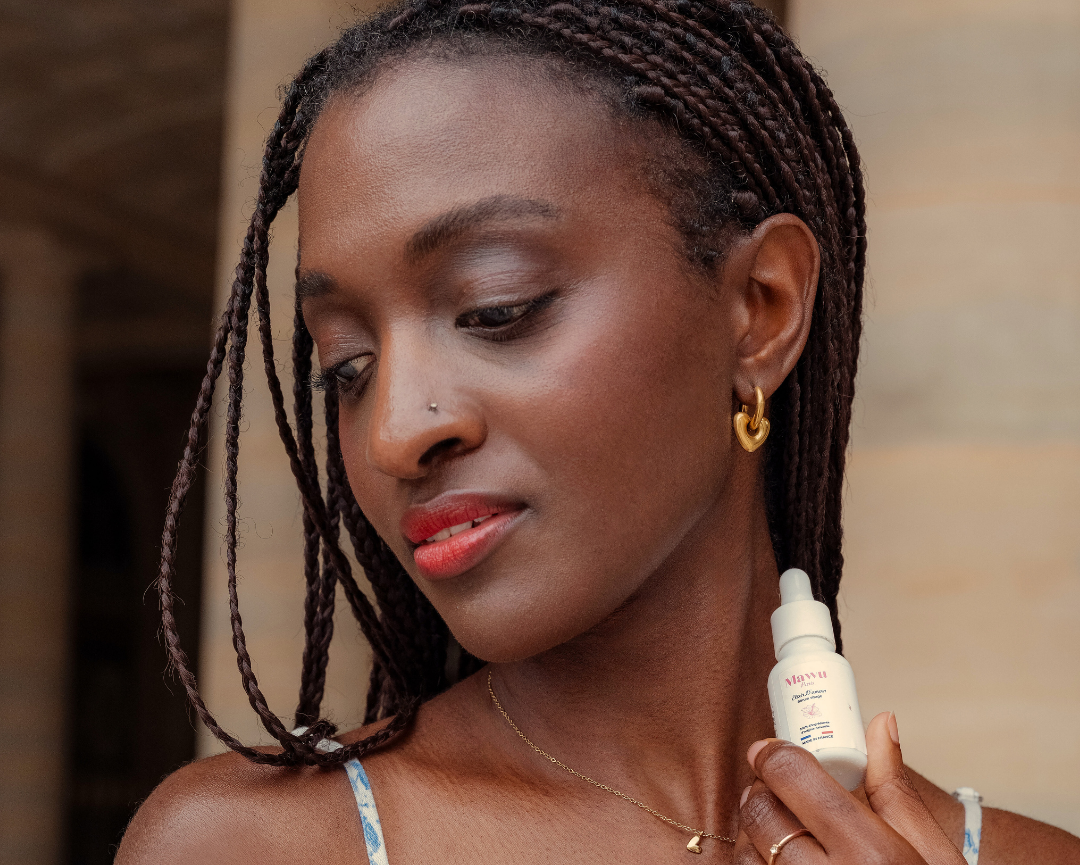 Belle femme noire, en robe à fleurs, avec de longues tresses et un sérum visage Élixir D'amour dans la main.