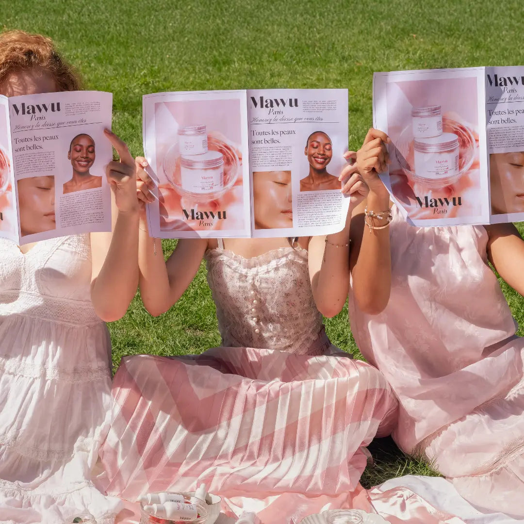 Trois filles à la peau claire avec des robes blanche et rose poudrée tenant le journal Mawu Paris devant leur visage.