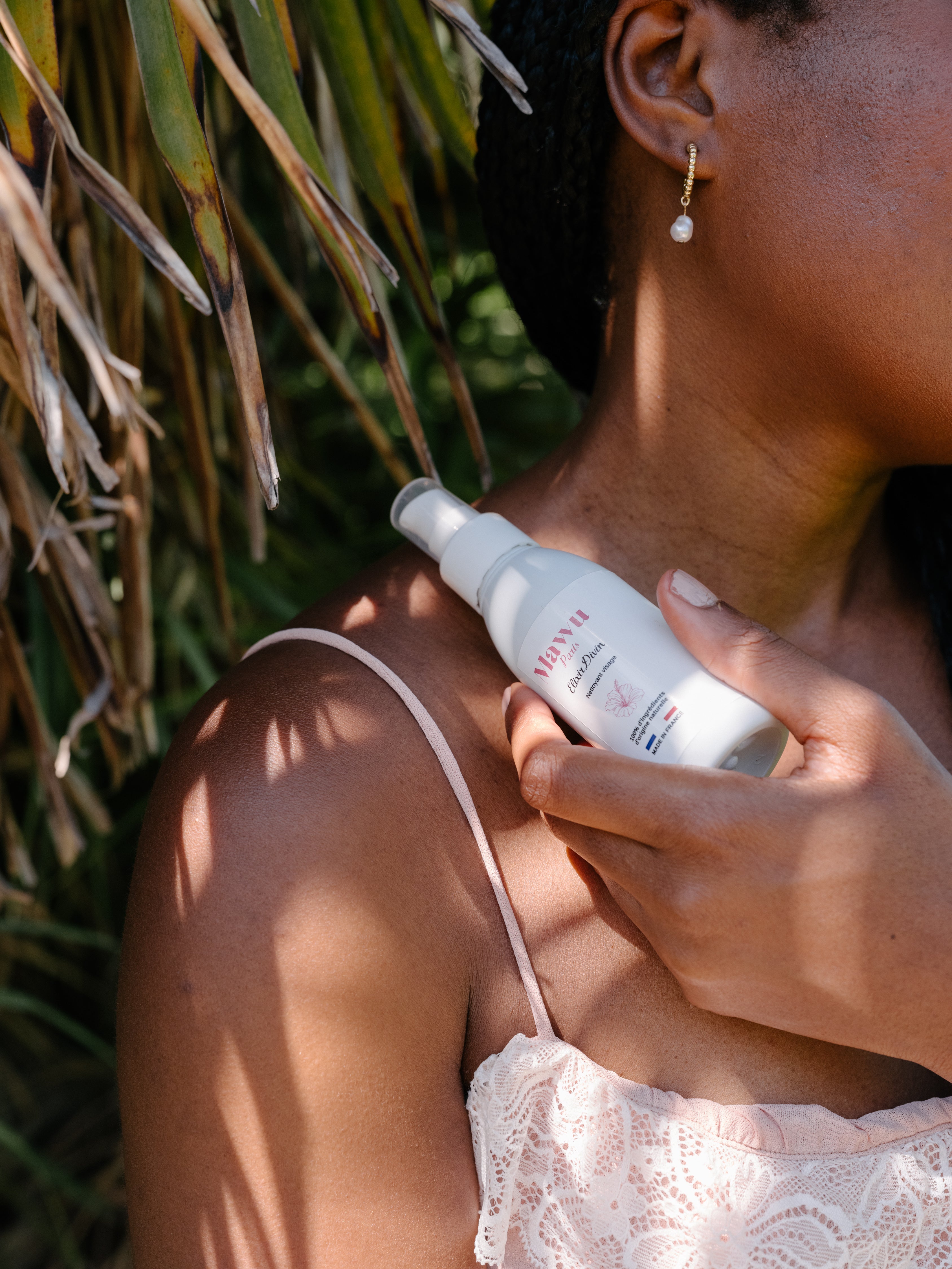 Une femme noire en débardeur rose poudré tient un flacon d'huile nettoyante Élixir Divin sur sa nuque.