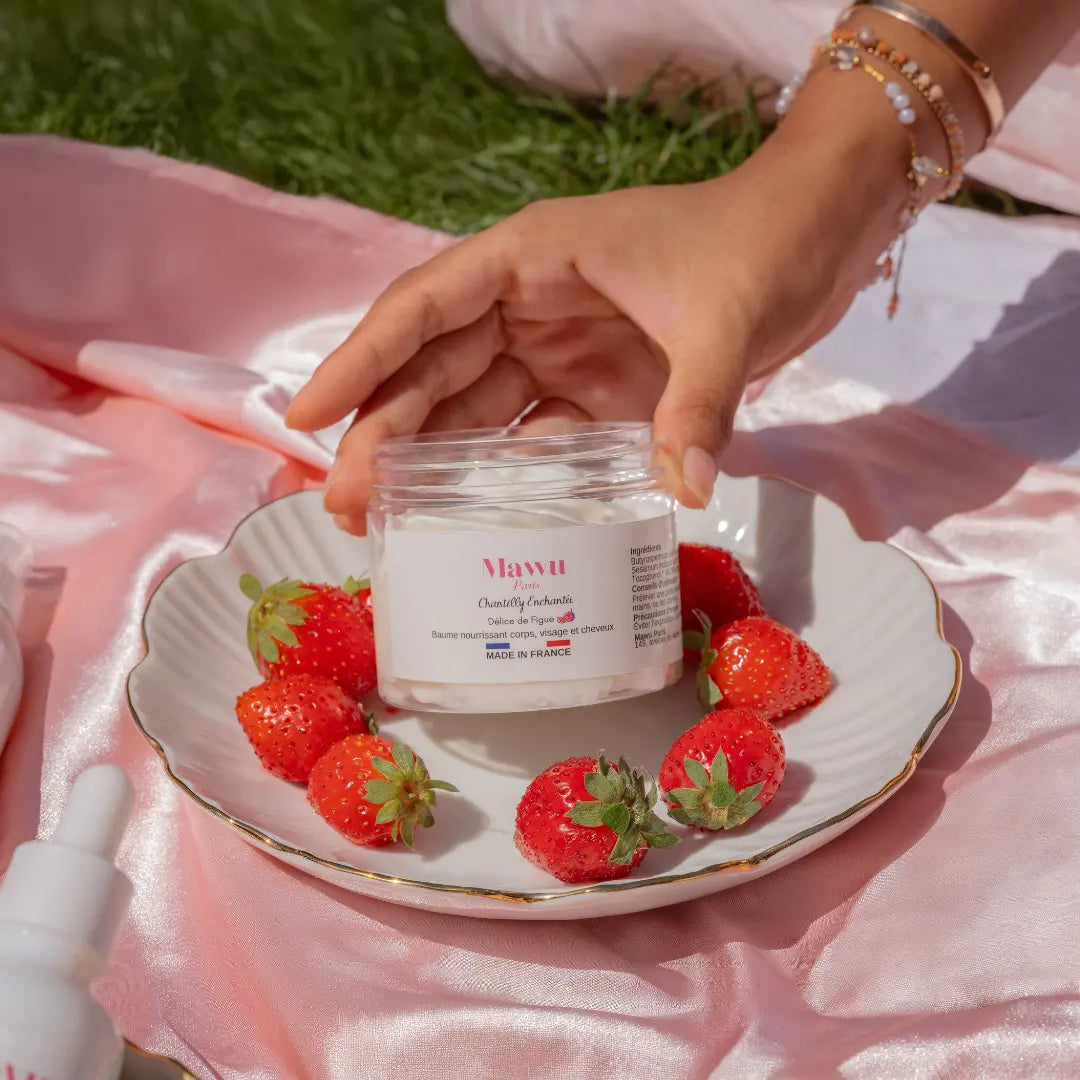 Une femme avec des bracelets au poigné tiens un peau de Chantilly Enchantée Mawu Paris, posé dans une assiette en porcelaine entourée de fraises fraîches. L'assiette es posée sur une nappe de satin rose au milieu de l'herbe.