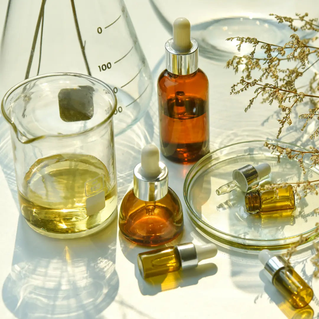 Sur une table blanche, une branche d'herbe, 3 minis flacons de sérum à pipettes blanches, 2 flacons ombrés à pipettes blanches, un verre bécher gradué, et un autre verre bécher contenant un liquide jaune.