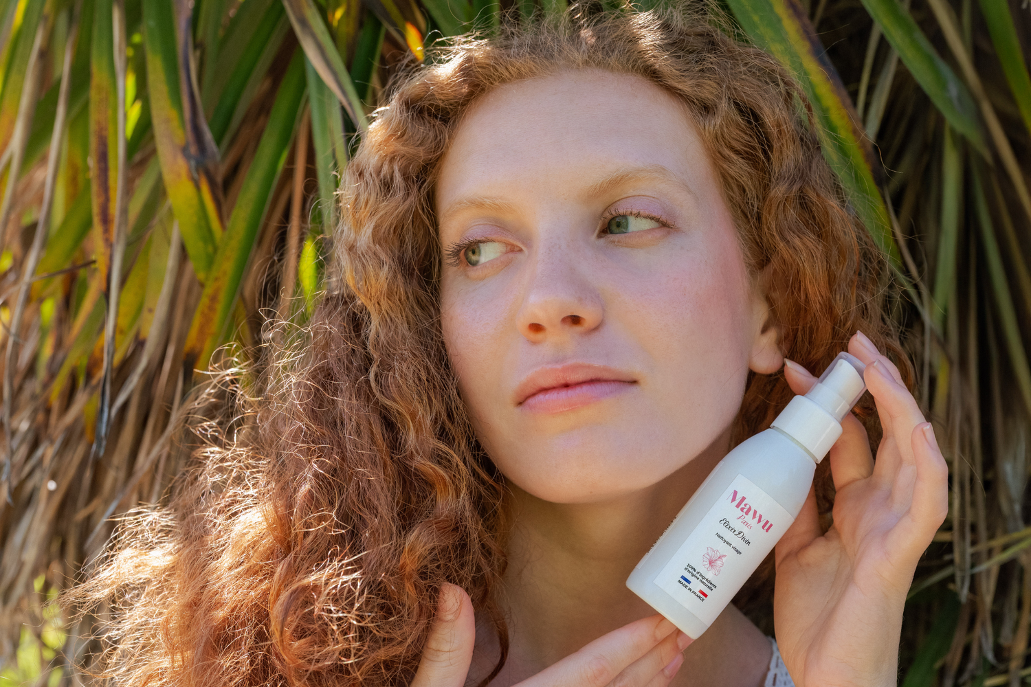 Jolie femme rousse à la peau claire yeux verts, face à un palmier tenant l'huile nettoyante Élixir Divin.
