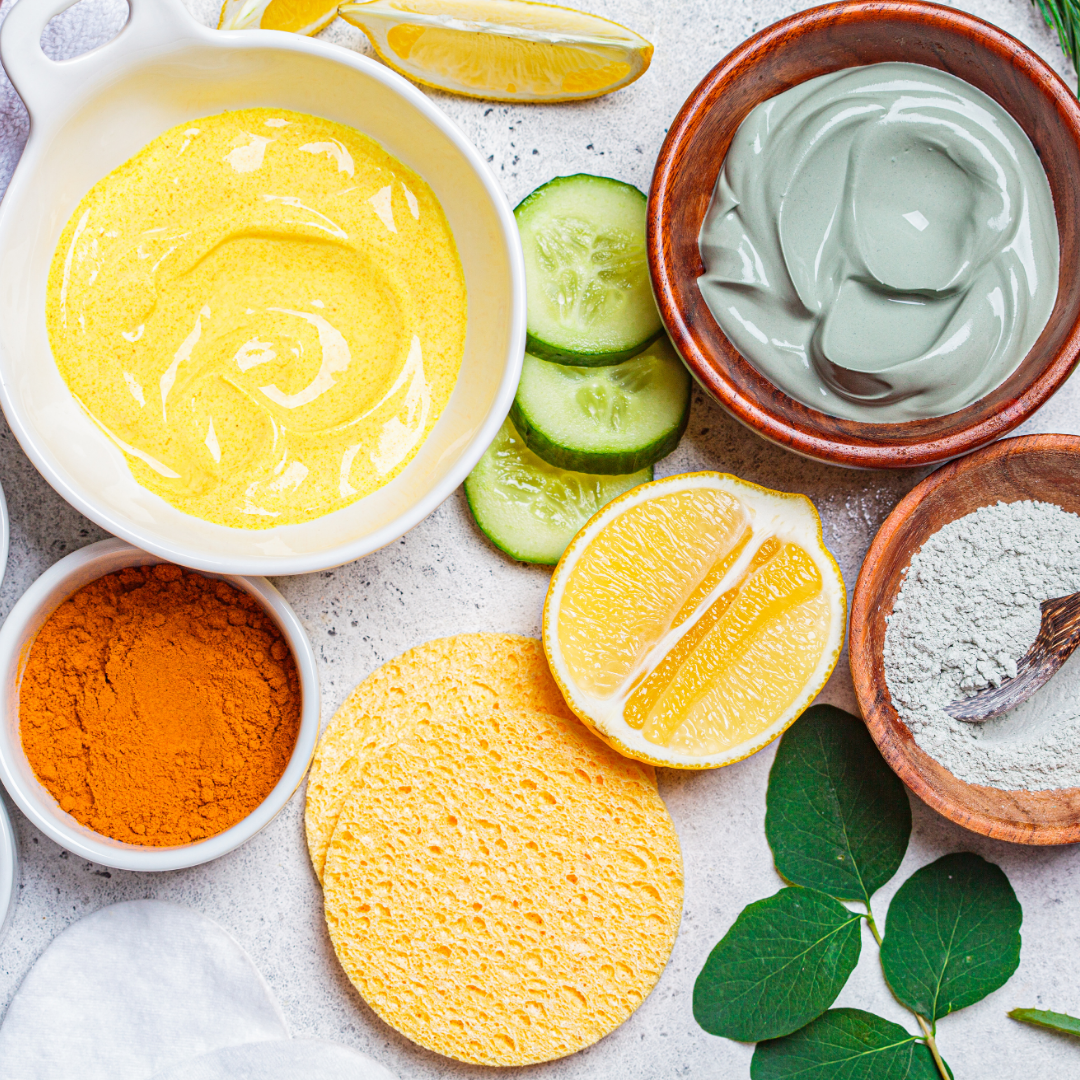 Sur une table en pierre gris clair, des morceaux de citron de concombre un ramequin porcelaine contenant du curcuma, un bol en bois contenant de l'argile blanche en poudre, un bol en porcelaine contenant un masque maison jaune, et un bol en bois contenant un masque à l'argile verte en pâte.