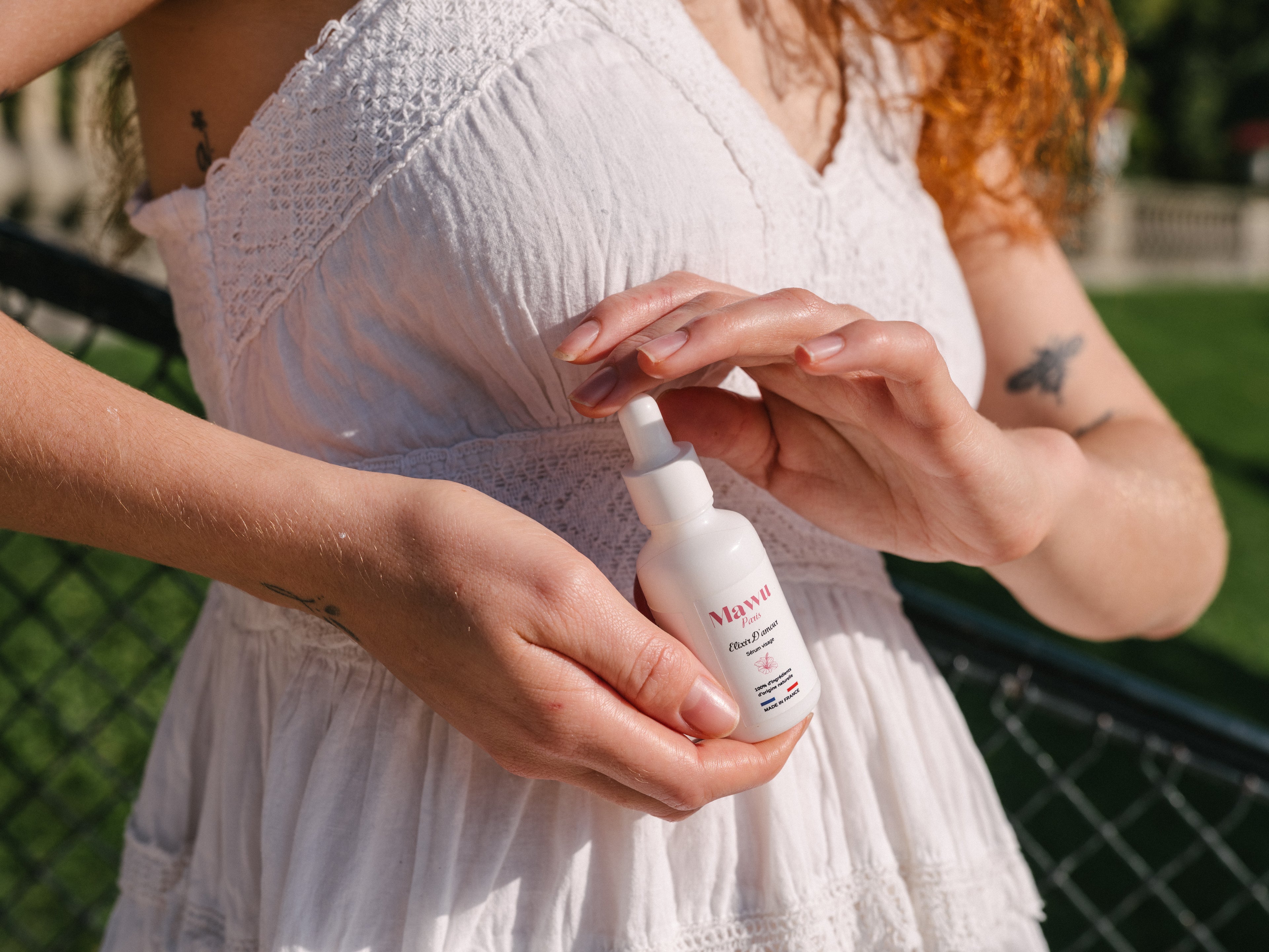 Une femme blanche aux cheveux roux porte une robe blanche, tient entre ses mains un flacon de sérum Élixir D'amour.