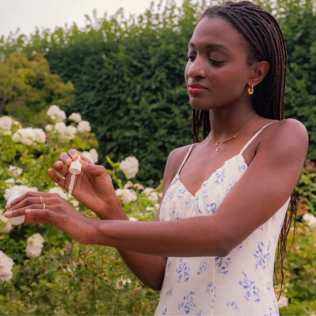 Une magnifique femme noire avec des tresses, une robe à fines bretelles, blanche à petites fleurs bleus, au milieu d'un jardin Français, emplit de rose blanches elle applique du sérum sur sa main gauche à l'aide d'une pipette blanche.