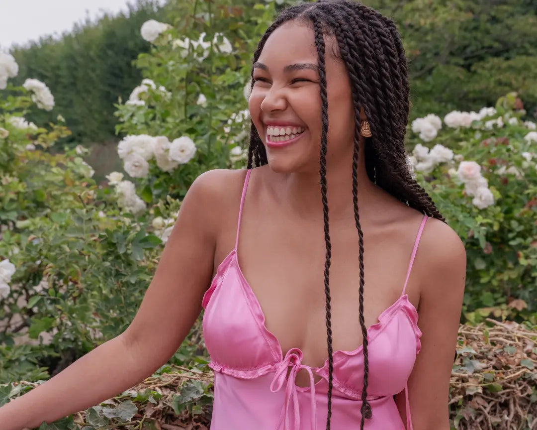 Une magnifique femme métisse souriante avec de longues tresses noirs, porte une longue robe rose à fines bretelles.