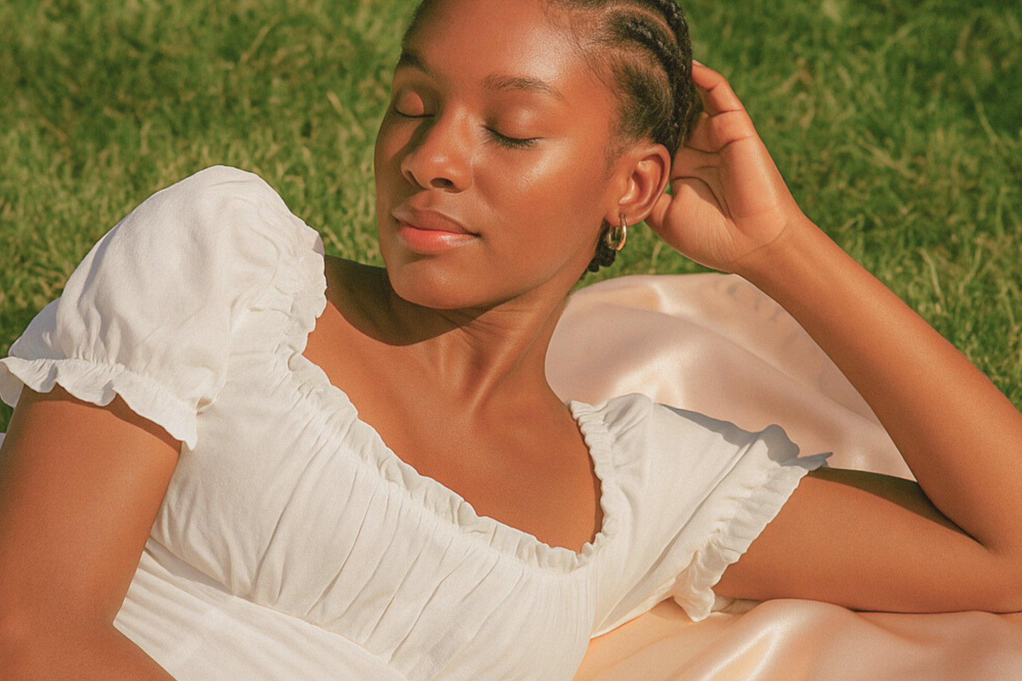 Une jolie femme noire dans un jardin français, allongée sur une nappe en satin rose. Elle à des tresses africaines et porte une robe blanche en coton, col bateau et manches courtes.