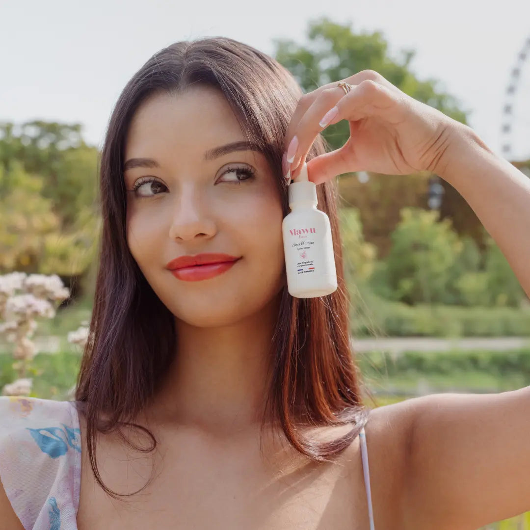 Une jolie femme aux longs cheveux noirs, sourie et tient un flacon de sérum Élixir D'amour contre sa joue gauche.