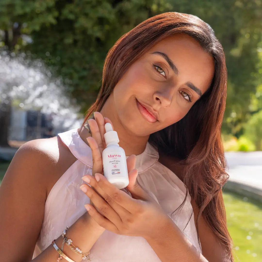 Jolie femme indienne à la peau mate et aux yeux verts debout face à une fontaine dans un jardin français, porte une robe rose poudré, tient un flacon de sérum visage Élixir D'amour dans les mains.
