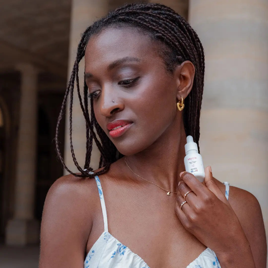 Une jolie femme noire avec de longues tresses, tient un flacon de sérum Élixir D'amour dans la main gauche.