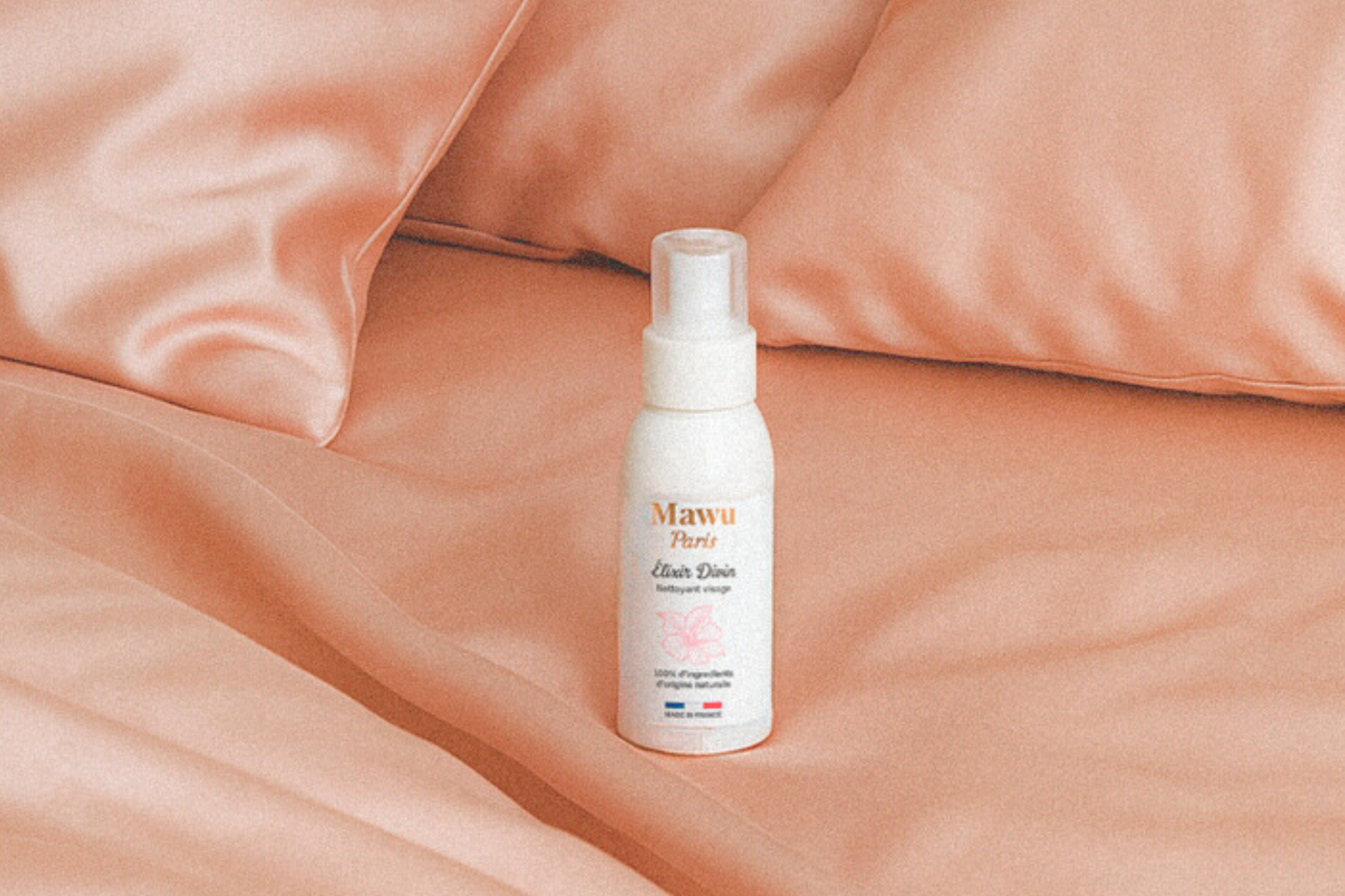 Huile nettoyante Élixir Divin, posée sur un lit avec housse et des taies d'oreiller en satin rose.