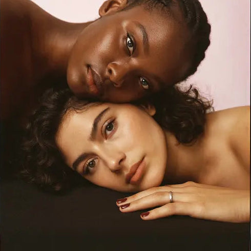 Une femme noire magnifique les cheveux tressés la tête posée, sur la tête d'ne  jolie femme à la peau mate et aux cheveux bouclés, aux ongles bordeaux avec un anneau à l'annulaire gauche, la tête posée sur un drap noir.