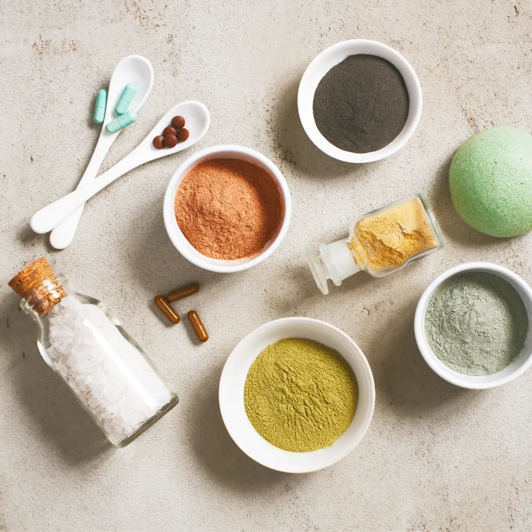 Sur une table en pierre gris clair, 4 bols en porcelaine blanche contiennent des des ingrédients naturels. Le premier du charbon en poudre, le deuxième de l'argile rouge, le troisième, de l'argile verte, le quatrième de l'argile blanche. Il y a une fiole en verre d'argile jaune, une fiole en verre de gros sel et deux cuillère en porcelaine contenant des compléments alimentaires.